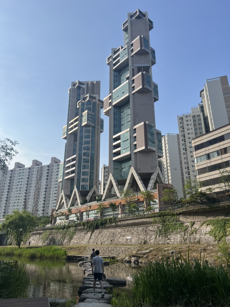 Two men cross a pedestrian stone bridge path across the Cheonggyecheon stream in Seoul, South Korea, with two large apartment buildings in the background.